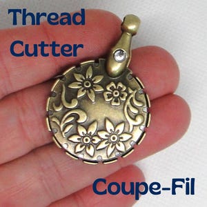 May include: A gold-toned thread cutter with a floral design and a clear rhinestone. The cutter is shaped like a circle with a small loop at the top. The text "Coupe-Fil" is printed below the cutter.