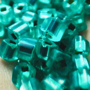 May include: Close-up of a collection of translucent, teal-coloured, cube-shaped beads. Each bead has a small, dark square within its centre. The beads are clustered together, creating a textured and shimmering effect.