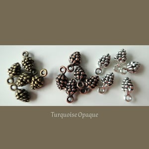 May include: Various metal pine cone charms in bronze, copper, and silver hues. Each charm features a loop for attaching to jewellery or crafts. The text "Turquoise Opaque" is visible at the bottom of the image.