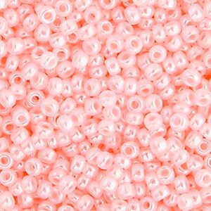 May include: A close-up shot of a pile of small, light pink seed beads. The beads are round with a hole through the center, and have a shiny, pearlescent finish. These beads are ideal for jewelry making and crafting.