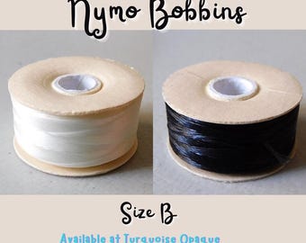 Może przedstawiać: Dwie szpulki nici, jedna biała, a druga czarna, na drewnianych szpulkach. Szpulki są oznaczone "Nymo Bobbins" i "Rozmiar B".