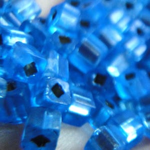 May include: A close-up shot of a collection of translucent blue beads. Each bead is a small, rectangular prism with a dark, square hole through its centre. The beads are scattered, creating a textured, shimmering effect.