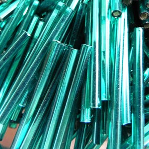 May include: A close-up of a pile of teal blue glass rods. The rods are smooth and shiny, and they are all different lengths.