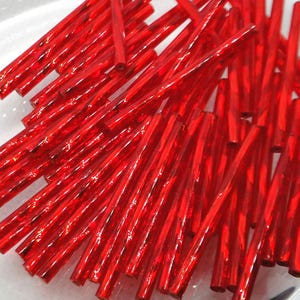 May include: A close-up shot displays a pile of vibrant red glass rods. The rods are cylindrical, with a glossy, transparent appearance. They are scattered across a white surface, catching the light and reflecting a bright, ruby-like hue. These glass rods are ideal for crafting.