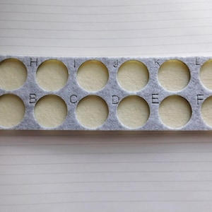 May include: A gray felt organizer with twelve circular cutouts, each containing a beige circle. Letters A through L are printed above and below the cutouts. The organizer is on a lined paper surface.