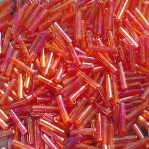 Peut inclure: Gros plan sur un tas de perles cylindriques oranges. Les perles ont une qualité translucide et irisée, reflétant la lumière avec des reflets roses et dorés. Elles sont dispersées au hasard, créant une surface texturée.