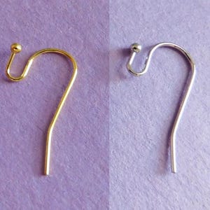 Può includere: Due ganci per orecchini, uno dorato e uno argentato, sono mostrati su uno sfondo viola. Ogni gancio ha una piccola sfera in cima e un design curvo per fissarsi all'orecchio.