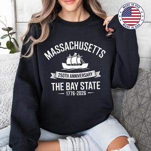 May include: A black sweatshirt with white text and a graphic of a sailboat. The text reads "MASSACHUSETTS", "250TH ANNIVERSARY", and "THE BAY STATE 1776-2026". The sweatshirt has a crew neck and long sleeves.