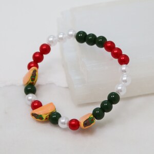Pode incluir: Um bracelete de contas coloridas com contas vermelhas, brancas e verdes. O bracelete tem três pequenos pingentes em forma de tacos.