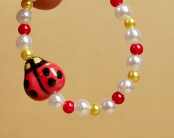 Könnte beinhalten: Ein Perlenarmband mit einem roten Marienkäfer-Anhänger mit schwarzen Punkten. Das Armband besteht aus weißen, roten und gelben Perlen. Der Marienkäfer-Anhänger ist rot mit schwarzen Details.