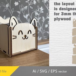 May include: A wooden cat-shaped box with a smiling face, ears, and a tail. The box is made of light-coloured wood and has a rectangular shape. Text on the right says "the layout is designed for 3mm thick plywood".