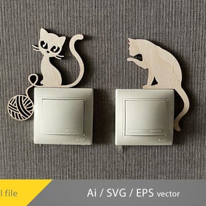 Op de afbeelding: Twee houten katvormige lichtschakelaars. Eén kat zit met een bol wol, de andere likt aan zijn poot. De lichtschakelaars zijn wit en de achtergrond is een grijze textuur.