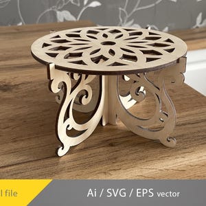 Puede incluir: Un pequeño soporte decorativo redondo de madera con un diseño floral cortado en la parte superior. El soporte tiene tres patas ornamentadas y está hecho de madera clara con un borde más oscuro. Las palabras "Digital file" y "Ai / SVG / EPS vector" están en la imagen.