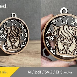 May include: Wooden Christmas ornament featuring a Highland cow design. The ornament is circular with a layered design, showcasing the cow's silhouette, snowflakes, and stars. The text "Tested!" is visible at the top. The ornament is made of light brown wood.