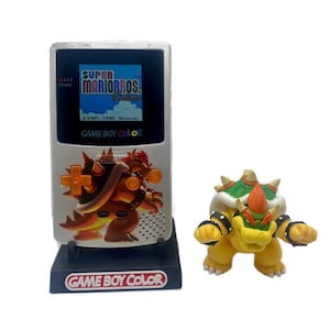 Op de afbeelding: Een zilveren Game Boy Color handheld-spelconsole met een standaard. De console heeft een scherm waarop het spel Super Mario Bros. Deluxe wordt weergegeven. Een klein plastic beeldje van Bowser, een personage uit de Super Mario Bros. videogame-serie, staat rechts van de console.