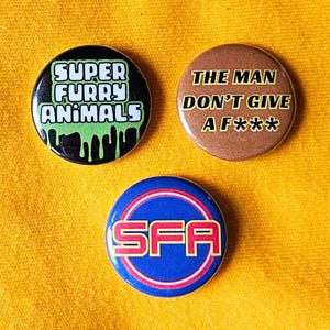 May include: Three circular pin badges with text. One black badge reads "SUPER FURRY ANIMALS" with green dripping accents. A brown badge says "THE MAN DON'T GIVE A F***". A blue badge has "SFA" in white.