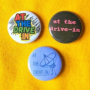 May include: Three round buttons with the text "AT THE DRIVE IN". One button is white with colorful text and scribbled lines. Another is dark with pink text. The third is blue with a satellite dish graphic.