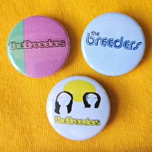 May include: Three round buttons featuring the band name "The Breeders" printed on them. The first button has a pink and green background with black text. The second button has a light blue background with blue text. The third button has a white background with a yellow sun and two black and white illustrations of women's faces. The text on the third button is in black.