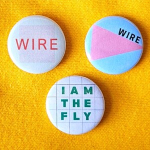 May include: Three round buttons with text and graphics. One button has the word "WIRE" in red. Another has a pink triangle on a blue background with the word "WIRE". The third button says "I AM THE FLY" in green.