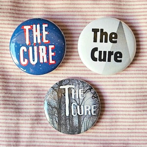 May include: Three round The Cure band badges. One badge is blue with white text, one is white with black text and a grey triangle, and one is a photo of trees with white text.