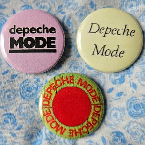 May include: Three round buttons with the text "Depeche Mode" in different styles. The first button is pink with black text, the second is yellow with black text, and the third is green with red and yellow text.
