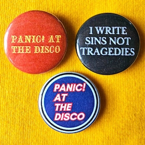 Peut inclure: Trois badges ronds avec des fonds noir, rouge et bleu. Les badges présentent le texte "Panic! At The Disco" en lettres blanches et jaunes. Le troisième badge porte le texte "I Write Sins Not Tragedies" en lettres blanches.