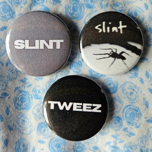 May include: Three round black and white buttons with band names. The first button has the word "SLINT" in white text on a gray background. The second button has the word "SLINT" in white text on a black background with a spider on a white background. The third button has the word "TWEEZ" in white text on a black background.