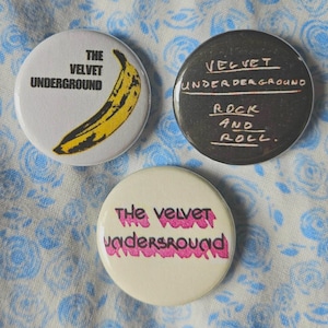 May include: Three round buttons with different designs featuring the band The Velvet Underground. The first button is white with a yellow banana and the text "The Velvet Underground". The second button is black with the text "Velvet Underground Rock and Roll". The third button is white with the text "The Velvet Underground" in pink.