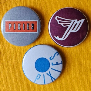May include: Three circular badges with various designs. One features the word "PIXIES" in black on a grey background with an orange rectangle. Another displays a white "P" with wings on a maroon background. The third has "PIXIES" in blue on a white background.