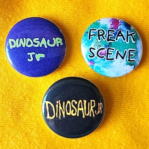 May include: Three circular badges. One is blue with "DINOSAUR JR" in green. Another is black with "DINOSAUR JR" in gold. The third badge is white with "FREAK SCENE" in black.