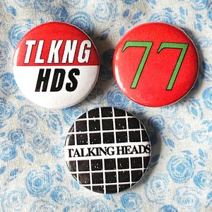 May include: Three round pinback buttons with Talking Heads imagery. The first button is red and white with the text "TLKNG HDS". The second button is red with the number "77" in green. The third button is black and white with a grid pattern and the text "TALKING HEADS".