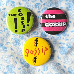 May include: Three round buttons with the word "GOSSIP" in different designs. One button is lime green with black text and exclamation points. Another is pink and black striped with white text. The third is yellow with red text and lightning bolts.