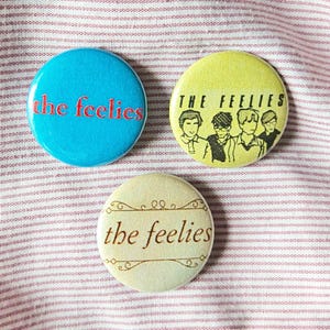 May include: Three round buttons with the text "the feelies" and band illustrations. One button is light blue with red text, another is yellow with a band illustration, and the third is off-white with brown text.