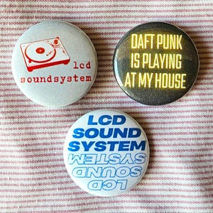 May include: Three round buttons with different designs. The first button is white with a red and black illustration of a record player and the text "lcd soundsystem". The second button is black with yellow text that reads "DAFT PUNK IS PLAYING AT MY HOUSE". The third button is white with blue text that reads "LCD SOUND SYSTEM" in a mirrored, upside-down design.