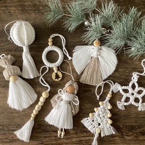 Op de afbeelding: Een verzameling handgemaakte macrame kerstversieringen in verschillende vormen, waaronder engelen, een krans, een sneeuwvlok en een kerstboom. De ornamenten zijn wit met houten kralen accenten en hangen aan witte koorden. Een takje groenblijvende planten voegt een feestelijke toets toe.