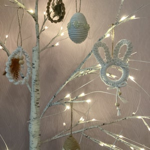 Puede incluir: Adornos de Pascua colgados de un abedul blanco iluminado con luces de hadas. Los adornos incluyen un conejito de macramé blanco, huevos tejidos y un huevo de fibra natural con adornos. El fondo es de color rosa pálido.