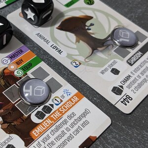 May include: Close-up of a board game setup with cards, dice, and tokens. The cards feature illustrations and text, including "ANIMAL LOYAL" and "EMILIEN, THE SCHOLAR." Two black dice with white symbols are present. Tokens with cube icons are also visible.