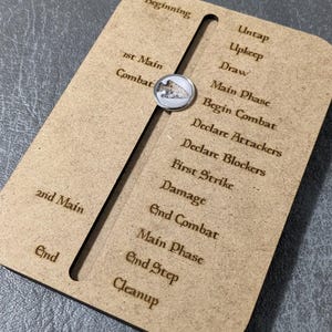 Könnte beinhalten: Ein Spielphasen-Tracker aus Holz mit einem Schiebemechanismus und graviertem Text. Der Text enthält Phasen wie "Beginn", "Kampf" und "Ende". Ein Metallschieber mit einem Vogelsymbol markiert die aktuelle Phase.