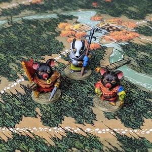 May include: Three small, hand-painted anthropomorphic animal figurines standing on a game board. One figure is a badger holding a spear, while the other two are mice, one carrying a red flag and a sword, and the other wearing colorful clothing.