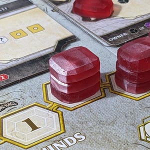 May include: A close-up of a board game in play. The game board features cards with text and illustrations, along with several red, gem-shaped game pieces. The word "ROUNDS" is visible on the board.