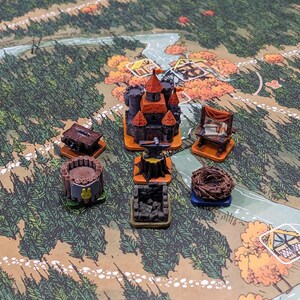 Peut inclure: Un ensemble de figurines de jeu de société miniatures comprenant un château central aux toits orange, un établi en bois, une structure circulaire en rondins avec une icône de lapin jaune, une forge en pierre avec une enclume, une table et un nid.