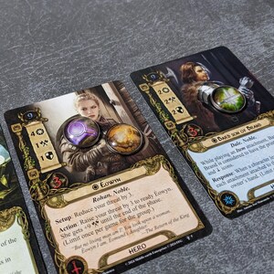 May include: Close-up of three trading cards featuring characters from a fantasy setting. The cards have detailed illustrations and text, with circular metal tokens on top. The cards are on a gray surface.