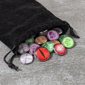 Può includere: Un sacchetto di velluto nero con coulisse riempito di gettoni rotondi in vetro cabochon. I gettoni presentano vari disegni colorati, tra cui un occhio di drago rosso, foglie verdi, pugni viola e simboli di spada bianchi su montature in metallo argentato.