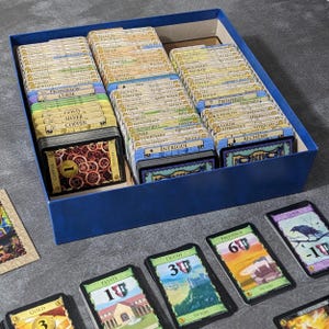 May include: A blue cardboard box filled with organized rows of rectangular game cards featuring various text labels and illustrations. Several individual cards are laid out in front of the box, including Estate, Duchy, Province, and Curse cards.