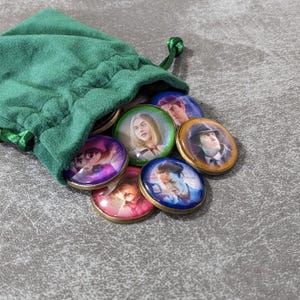 May include: A green velvet drawstring bag with several round, gold-rimmed tokens spilling out. Each token features a different portrait of a person. The tokens are various colours, including pink, blue, and purple.