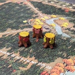 Peut inclure: Trois souches d'arbres miniatures marron avec des sommets jaunes, chacune ornée de petites icônes en forme d'animaux sur les côtés. Une souche porte des renards rouges, une des souris roses et une des lapins jaunes, posées sur un plateau de jeu.