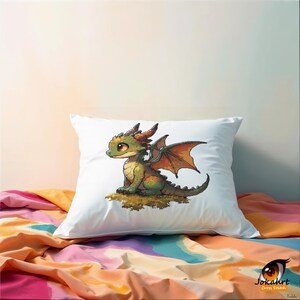 Pu&ograve; includere: Un cuscino quadrato bianco con un'illustrazione stampata di un piccolo drago verde con ali arancioni e una coda spinosa. Il drago &egrave; seduto su un pezzo di terra marrone. Il testo Jokahrt Cross Stitch &egrave; visibile nell'angolo in basso.
