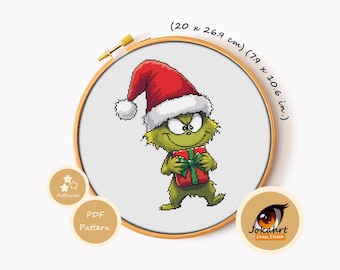 May include: A cross stitch pattern in a wooden hoop featuring a green creature wearing a red Santa hat and holding a red gift with a green ribbon. The text reads 20 x 26.9 cm and 7.9 x 10.6 in. Includes a logo with the text Jokahrt Cross Stitch.