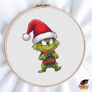 May include: A cross-stitch embroidery in a wooden hoop depicts a green cartoon character wearing a Santa hat and holding a red gift box with a green bow. The character has a mischievous grin. The background is white.