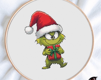 May include: A cross-stitch embroidery in a wooden hoop features a green cartoon character wearing a Santa hat and holding a red gift box with a green bow. The character has a mischievous grin. The background is white.